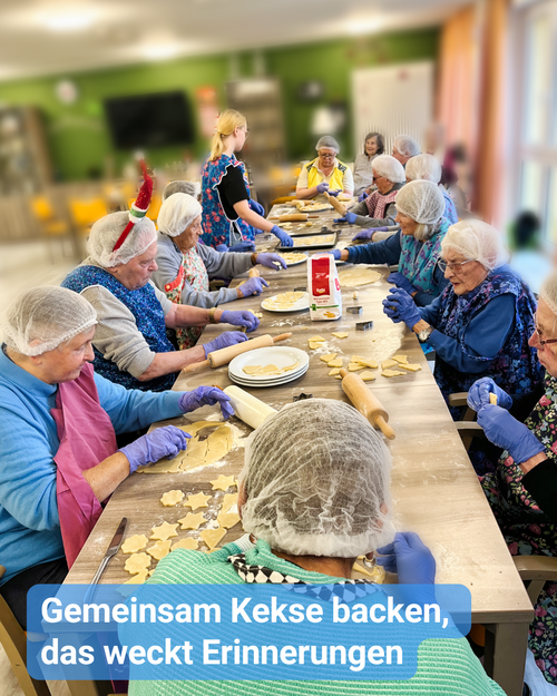 Senioren und Senioren sitzen gemeinsam an einem langen Tisch und stechen Weihnachtsplätzchen aus. Bildbeschriftung: „Gemeinsam Kekse backen, das weckt Erinnerungen”
