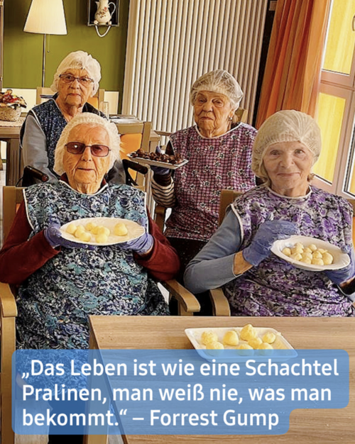 Vier betagte Frauen, jede mit einer Hygienehaube auf dem Kopf, sitzen um einen Tisch in dem, was offenbar ein gemeinschaftlicher Speiseraum eines Seniorenheims ist. Sie teilen sich Teller mit kleinen, hell gefärbten Süßigkeiten oder Konfekten, möglicherweise Pralinen. Vor jeder Frau steht ein Teller mit diesen Köstlichkeiten.  Eine Frau befindet sich im Vordergrund, am nächsten zum Betrachter. Die Frauen lächeln sanft und scheinen sich aneinander und an der gemeinsamen Zeit zu erfreuen. Die Umgebung ist schlicht und sauber, geprägt von sanften Farben und warmem Licht.  Ein Zitat aus Forrest Gump ist am unteren Rand zu sehen: "Das Leben ist wie eine Schachtel Pralinen, man weiß nie, was man bekommt."